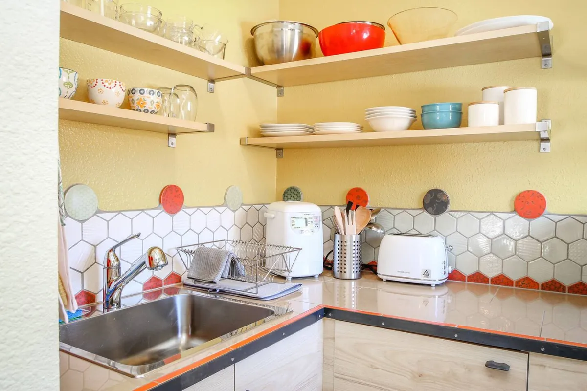 The image depicts a cheerful, well-equipped kitchen scene. Prominent features include a sink, open shelves stocked with colorful bowls and kitchen utensils, and a toaster, all bathed in natural light. The arrangement suggests a space ready for cooking, baking, or socializing over a homemade meal.