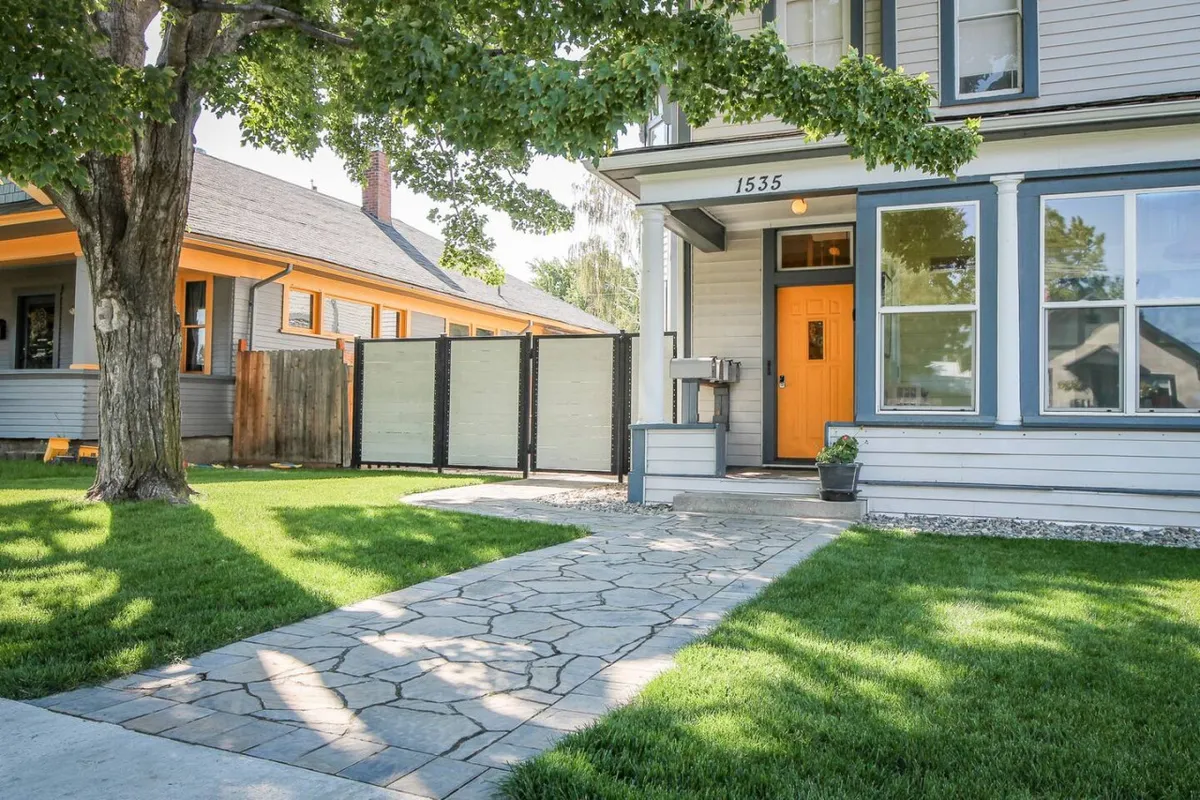 Shows the front of the property with a vibrant orange door, a lush green lawn, mature tree, and a textured stone pathway. The scene invites outdoor relaxation and hints at gatherings, barbecues, and time spent with pets or loved ones on the lawn.