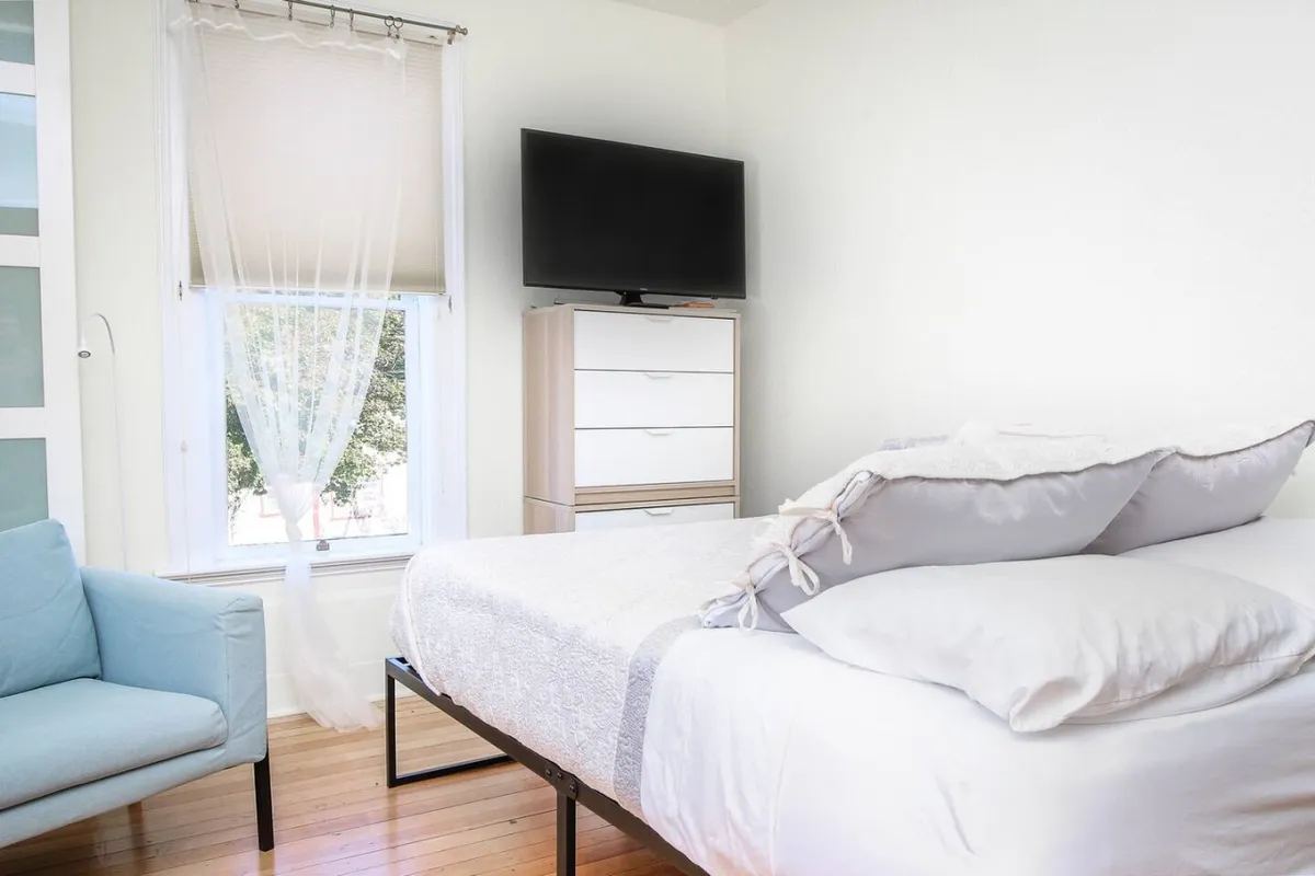 A bright, cozy studio space featuring a queen-sized bed with soft pillows, a stylish blue armchair, a large smart television, and a window adorned with sheer curtains that lets in abundant natural light. The arrangement subtly showcases the sitting area and entertainment amenities.