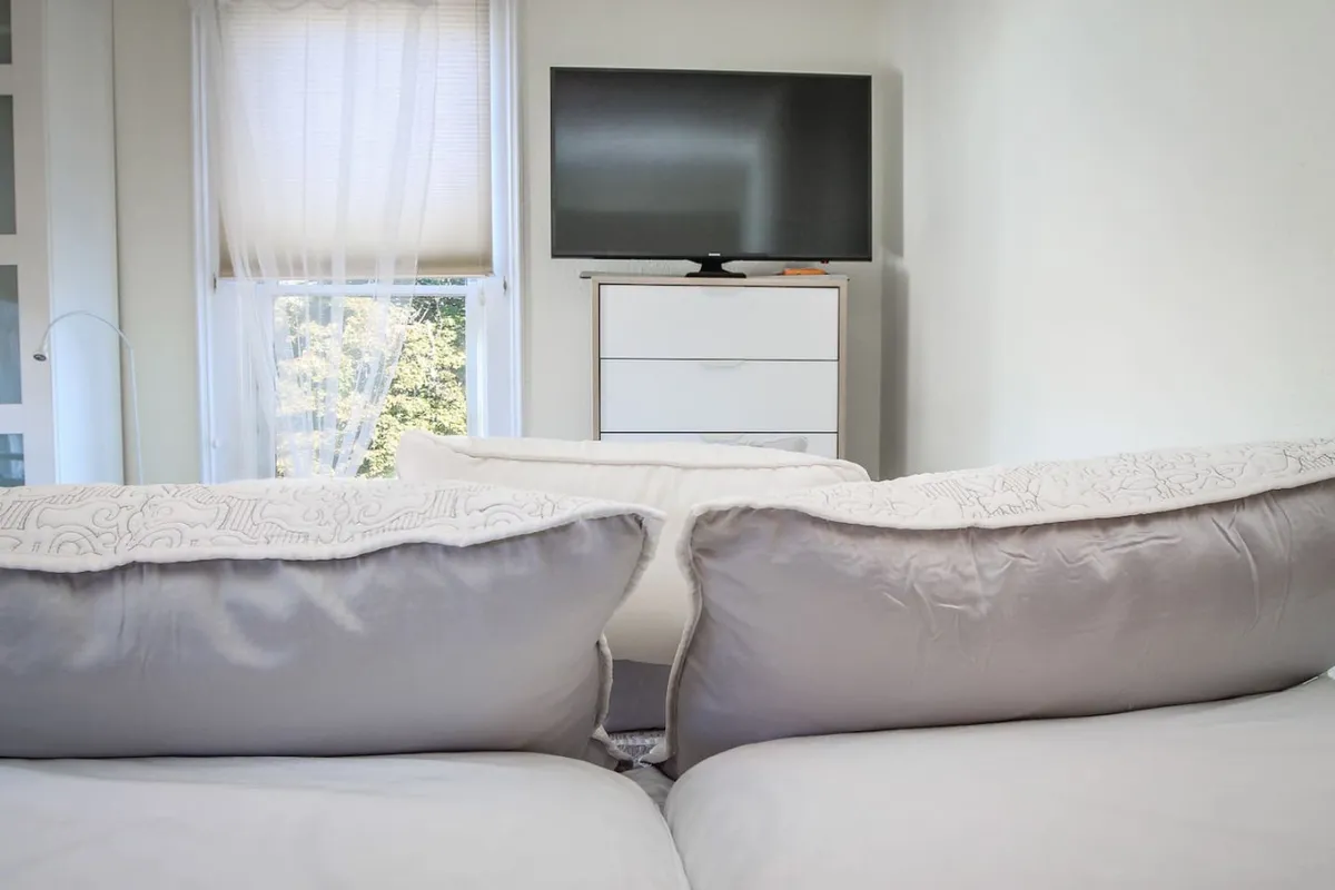 A serene sitting area designed for relaxation, featuring oversized pillows, a television set for entertainment, and a window with sheer curtains that allow gentle daylight to enter, creating a soothing and inviting atmosphere.