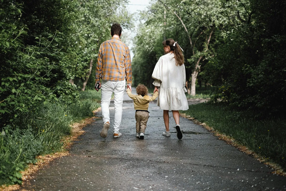 A joyful family strolls along a picturesque path lined with green trees, with a child holding hands with both parents. The lush surroundings evoke a sense of exploration, connection, and access to nearby parks, reinforcing the walkable neighborhood theme.