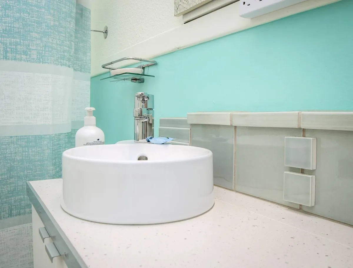 A bright and inviting bathroom scene, highlighting a modern round sink, sleek faucet, aqua-colored walls, and a textured shower curtain. Plush towels and countertop space are visible, suggesting thoughtful attention to guest comfort and personal care routines.