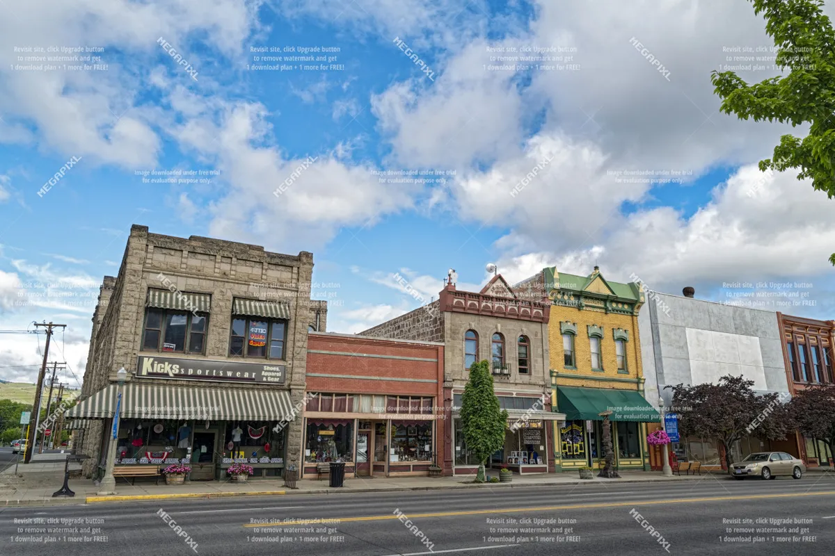A charming streetscape featuring historic buildings, unique storefronts, and leafy street trees lining the sidewalk. People can be seen strolling, shopping, and enjoying the vibrant, walkable atmosphere of Baker City's downtown area.