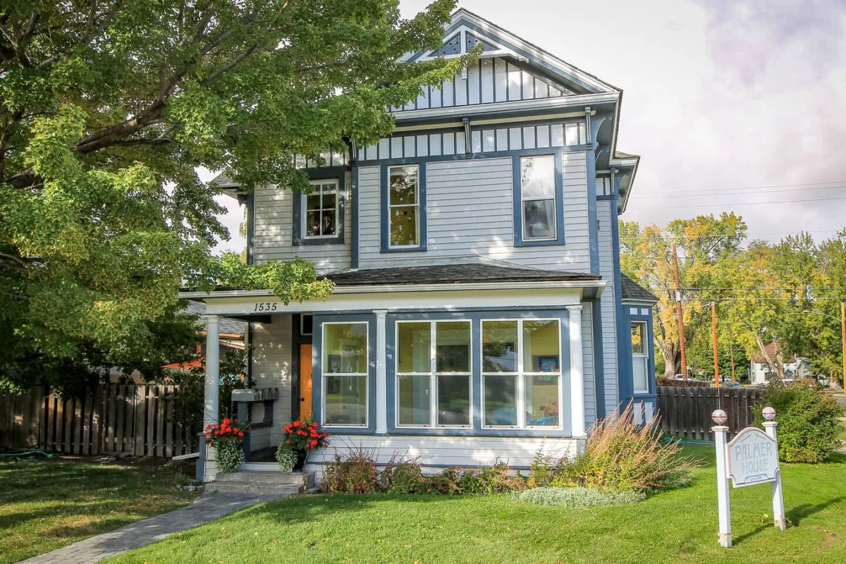 The inviting exterior of a classic blue Victorian house with large windows and a welcoming front porch, set against a sunset backdrop. Signposts and garden activities hint at friendly neighborhood living and leisurely exploration.