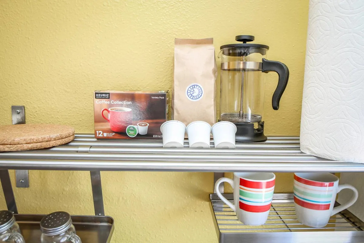 The image displays a cozy coffee station, featuring a coffee maker, a selection of coffee pods (including Sorbenots, as described), colorful mugs, a coffee bag, and a cork coaster on the kitchen countertop. The station is lit warmly, inviting guests to start their day or relax with a hot beverage.